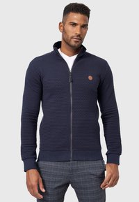 Marineblå zip-jakke med tekstureret stof, ribstrik ved håndleddene og kanten, samt et lille logo-patch på brystet. Båret over en hvid T-shirt.