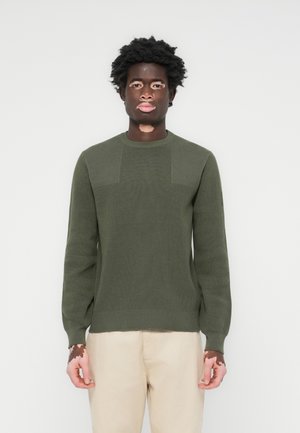 Olive green knitted sweater with long sleeves, ribbed texture, and panel details on the front. Paired with light beige pants.