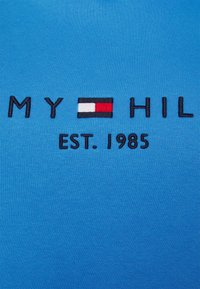 Tela azul con texto bordado "MY HIL" rodeando un rectángulo rojo, blanco y negro, y "EST. 1985" abajo.