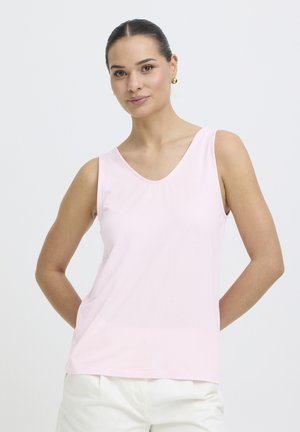 Femme aux cheveux foncés attachés, portant des boucles d'oreilles dorées, un haut sans manches rose clair et un pantalon blanc, debout devant un fond blanc.