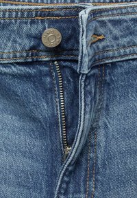 Denim-farkut vetoketjulla, sininen väri, pronssinappi logolla, kontrastisaumaus ja näkyvä kangasrakenne.