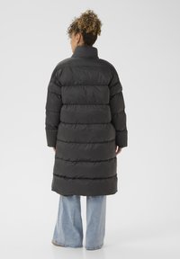 Cappotto imbottito nero con colletto alto, caratterizzato da un design trapuntato, maniche lunghe e una silhouette fino al ginocchio, abbinato a jeans svasati azzurri chiari.