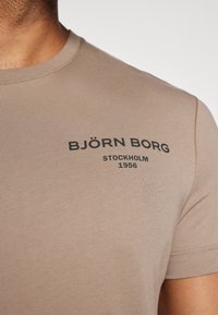 Beige bomull t-skjorte med rund hals og korte ermer. Har svart trykket tekst: "BJÖRN BORG STOCKHOLM 1956" på forsiden.