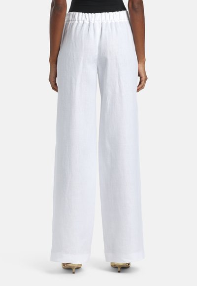Femme portant un pantalon blanc en lin taille haute et jambes larges, ainsi que des escarpins beiges à petit talon, vue de dos sur un fond uni.