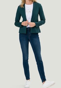 Tyrkysový blazer s upraveným střihem, na jedné knoflíkové zapínání, v kombinaci s bílým grafickým tričkem a tmavě modrými džíny, doplněno bílými teniskami.