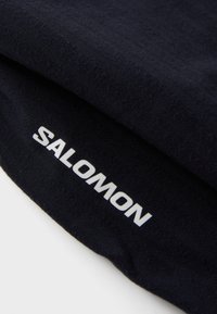 Juodas audinys su lygia tekstūra; krašte ryškiai matomas baltas "SALOMON" logotipas.