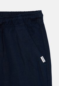 Námořnické modré kalhoty s elastickým pasem, bočními kapsami a malou značkovou visačkou. Materiál se zdá být lehký s hladkou texturou.