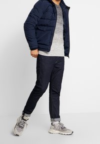 Marinblå puffjacka, grå stickad tröja, mörka jeans, ljusgrå sneakers med meshdetaljer, står på en vit bakgrund.