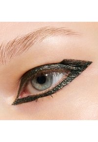 Eyeliner vert métallique avec un design ailé net autour d'un œil bleu. La peau présente une texture lisse et légèrement réfléchissante. Sourcil subtil au-dessus.