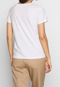 White cotton t-shirt with a classic crew neckline and short sleeves, featuring a smooth texture and relaxed fit, paired with tan trousers.