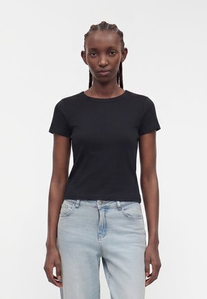 Ung kvinde med flettet hår iført en tætsiddende sort t-shirt med korte ærmer og lyseblå højtaljede jeans, stående foran en ensfarvet baggrund.