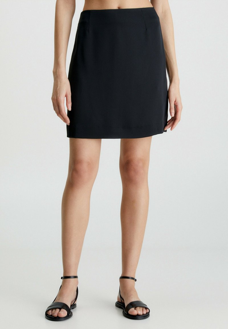 Mini-jupe noire à texture lisse, coupe droite, poches latérales et ourlet net, portée avec des sandales plates noires.