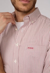 Chemise rayée rouge et blanche avec un col boutonné et une poche poitrine, ornée de subtils accents brodés au-dessus de la poche.