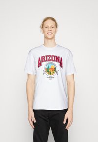 Camiseta de algodón blanca con "ARIZONA" en letras rojas y un gráfico colorido de un paisaje desértico con cactus, que presenta acentos de calaveras de vaca.