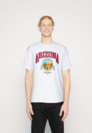 Camiseta de algodón blanca con "ARIZONA" en letras rojas y un gráfico colorido de un paisaje desértico con cactus, que presenta acentos de calaveras de vaca.