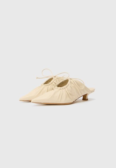 Mules en cuir beige avec des détails froncés et un design à bout pointu. Talon bas et fines lanières pour un ajustement réglable. Texture lisse.