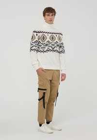 Sweter z golfem w kolorze białym ozdobiony wielokolorowymi wzorami geometrycznymi, połączony z khaki spodniami cargo z czarnymi akcentami i kieszeniami.