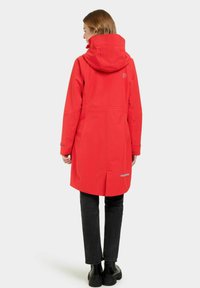 Manteau imperméable rouge avec capuche, design allongé et détails réfléchissants. Présente une texture lisse et des poches latérales. Porté avec un pantalon noir.