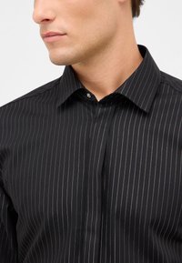 Mann in einem schwarzen gestreiften Hemd mit weitem Kragen, von der Halsmitte bis zur Mitte des Oberkörpers zu sehen, von links blickend.