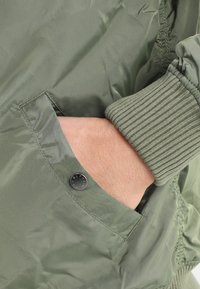 Veste bomber vert olive avec des poignets côtelés et une poche dotée d'un bouton-pression en métal noir. Texture lisse et tissu léger.
