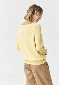 Pulover galben din material moale, cu guler rotund și tiv cu nervuri. Purtați cu pantaloni bej deschis, care au o croială lejeră.