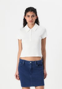 Vit bomullspolo med spetsig krage, korta ärmar och en kort design, ihop med en blå denim minikjol med fickor.