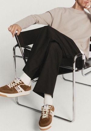 Sneaker di suede marrone con accenti bianchi, lacci crema e suola in gomma. Il modello indossa pantaloni neri e un sudore beige chiaro, seduto su una sedia.