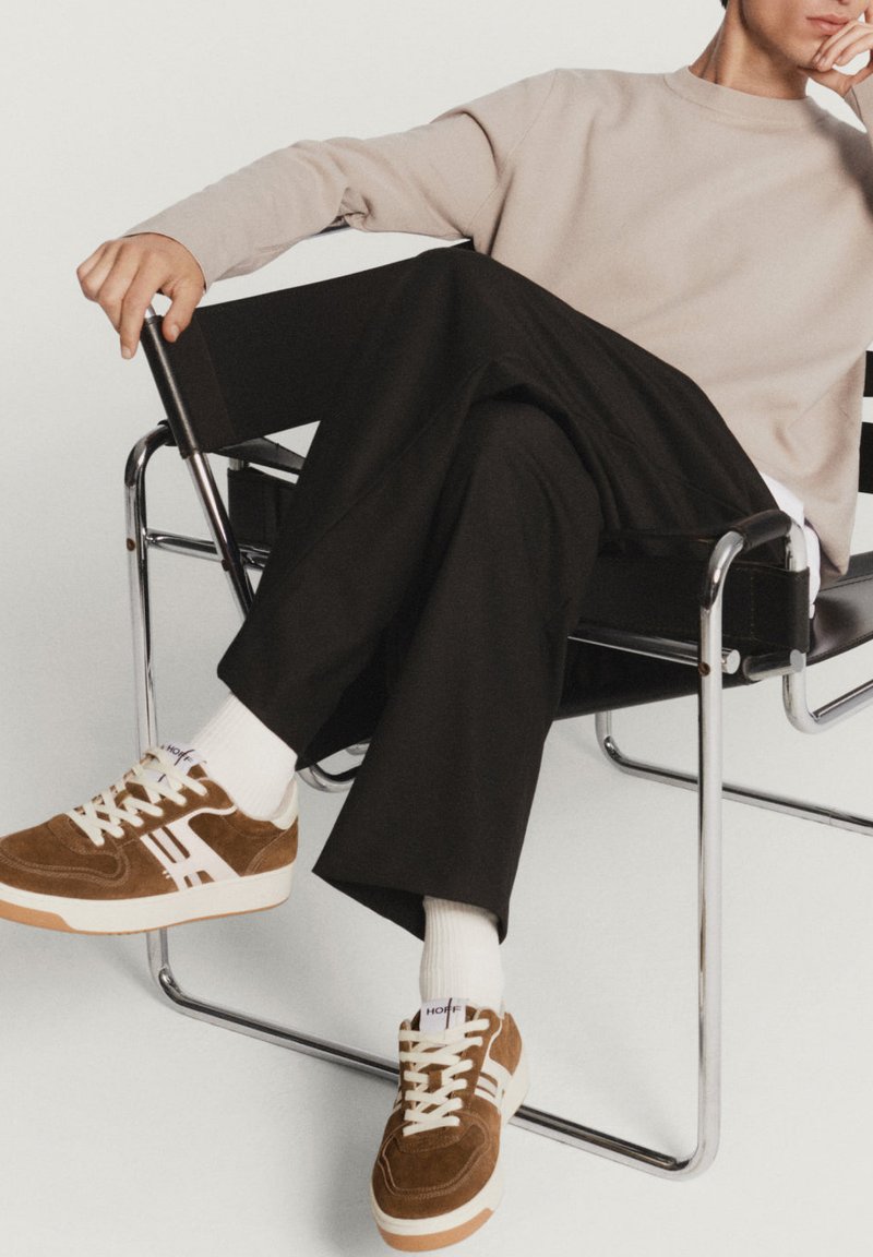 Sneaker di suede marrone con accenti bianchi, lacci crema e suola in gomma. Il modello indossa pantaloni neri e un sudore beige chiaro, seduto su una sedia.
