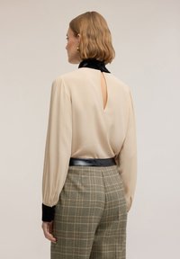 Beżowa bluzka z czarnym aksamitnym kołnierzykiem i dekoltem w kształcie łezki, zestawiona z kratkowymi spodniami w odcieniach zieleni i beżu oraz czarnym paskiem.