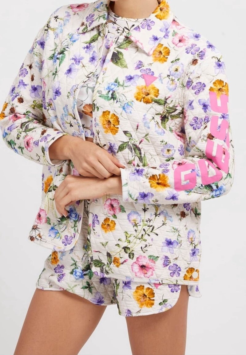 Veste matelassée à motif floral de couleur crème, avec un logo rose sur la manche. Associée à un short assorti à motifs floraux.