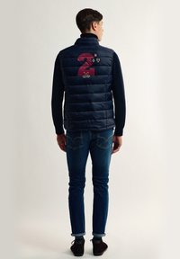 Chaleco de plumas azul marino con un patrón de acolchado horizontal, presenta un gran número "2" burdeos y parches en la parte posterior, combinado con jeans azules.