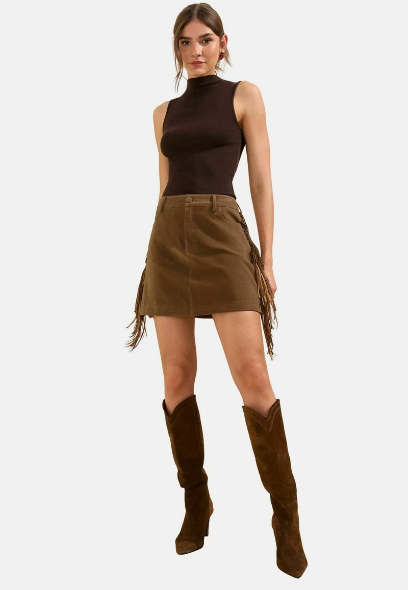 Woman wearing sleeveless brown top, brown suede skirt with fringe details, and knee-high brown suede boots standing against plain background.