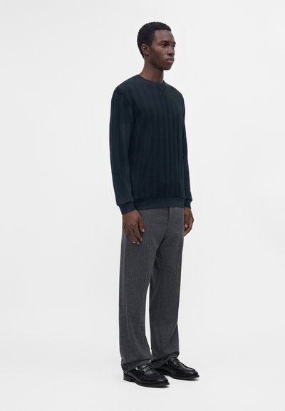 Un homme se tient de profil droit, portant une chemise à manches longues texturée foncée, un pantalon gris et des chaussures habillées noires sur fond blanc.
