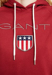 Sudadera con capucha roja con cordones blancos que presenta un logotipo gris de "GANT" y un parche en forma de escudo con franjas rojas y azules y tres estrellas blancas.