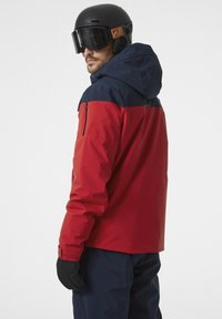 Veste de ski rouge et marine avec capuche, poche zippée à l'avant, poignets ajustables, portée avec des gants noirs et un casque noir avec lunettes de ski.