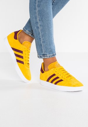 Person iført klare gule ruskindssneakers med lilla striber og hvide såler, kombineret med opbukket blå jeans på et hvidt gulv.