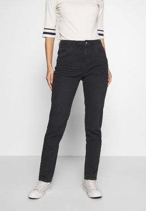 Osoba ubrana w czarne, wysokie, dopasowane jeansy, biały top z krótkim rękawem i czarnymi paskami na rękawach oraz białe trampki, stojąca na szarej podłodze.