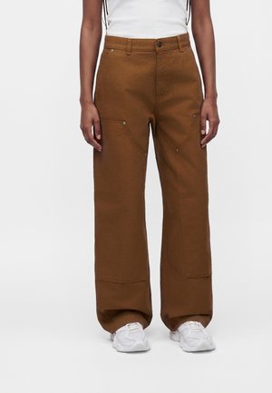 Personne portant un pantalon marron ample avec des poches plaquées devant et des baskets blanches, montrée de la taille aux pieds sur un fond uni.