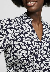 Blouse bleu marine à motif floral blanc, dotée d'un col en V et de manches courtes bouffantes. Fabriquée en tissu léger avec une texture lisse.