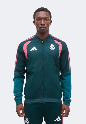 Bărbat purtând o jachetă sport de culoare teal închis și pantaloni cu dungi roz și siglele Real Madrid și Adidas, stând cu fața înainte.
