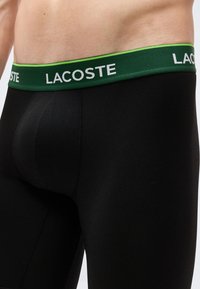 Κοντινό πλάνο ενός ατόμου που φοράει μαύρα ανδρικά μπόξερ Lacoste με πράσινη μέση που φέρει το λογότυπο της Lacoste με λευκά γράμματα.