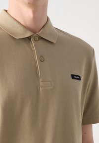 Homme portant un polo beige à manches courtes avec deux boutons attachés et un petit logo rectangulaire noir Calvin Klein sur la poitrine gauche.