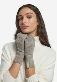 Grå stickade handskar med ribbad design och dekorativa pärldetaljer. Har tygetiketter på muddar. Modellen bär en ljus tröja.