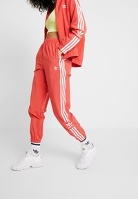 Červená tepláková souprava s bílými pruhy, vybavená elastickým pasem a manžetami u kotníků. Kombinována s bílými teniskami a pruhovanými ponožkami.