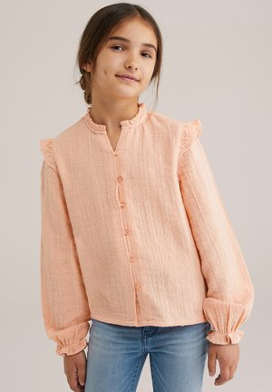 Junges Mädchen mit dunklen Haaren in einer peachfarbenen, strukturierten Bluse mit Rüschen und blauen Jeans, das leicht lächelt vor einem einfarbigen Hintergrund.