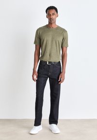 Grünes kurzärmliges T-Shirt, dunkle Jeans und weiße Sneakers. Lässiger Schnitt, strukturierter Stoff und eine metallische Gürtelschnalle als bemerkenswerte Details.
