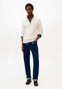Giovane uomo in piedi con una mano in tasca, indossa un maglione bianco a trecce sopra una camicia di denim blu, jeans scuri e scarpe nere.