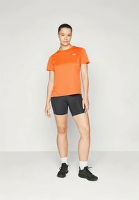 Oranžová športová košeľa s krátkym rukávom v kombinácii s čiernymi športovými šortkami. Model má na sebe čierne tenisky a biele členkové ponožky. Jednoduchý dizajn, bez vzorov.