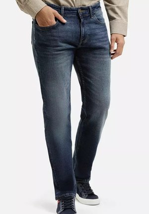 Mężczyzna w ciemnoniebieskich dżinsach, beżowej koszuli i czarnych sneakersach, z lewą ręką w kieszeni, stojący na białym tle.