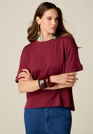 Femme aux longs cheveux bruns portant une chemise bordeaux à manches courtes, un jean bleu, de grands bracelets en bois et de longues boucles d'oreilles vertes et marron.